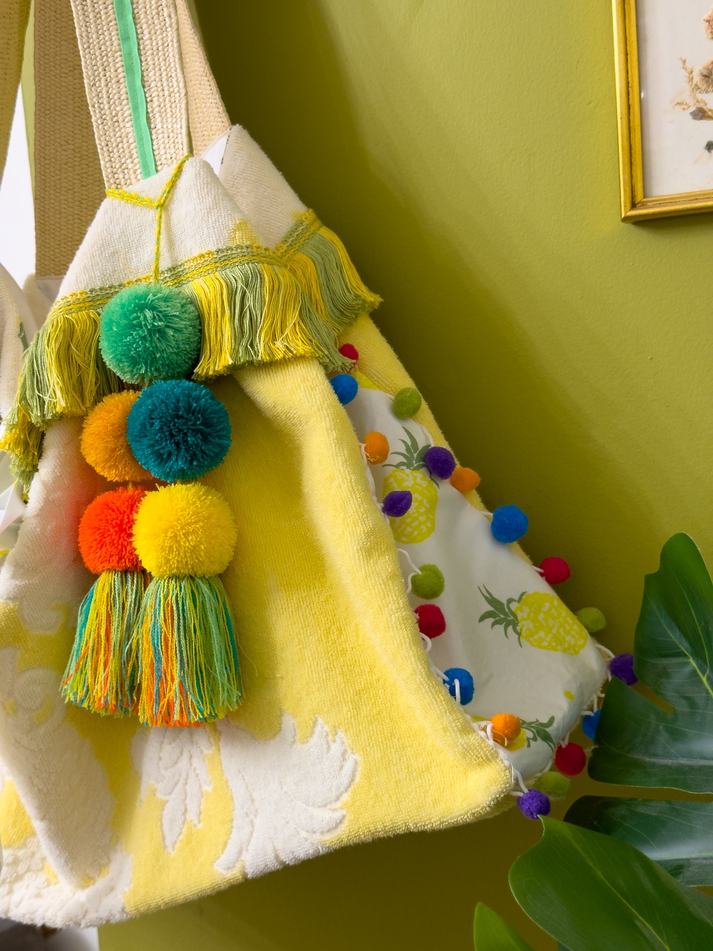 Close up photo of the side of a beach tote bag made from a vintage yellow and white towel, the side of the bag has a white with pineapple pattern and rainbow pom pom trim, the top edge of the bag has a yellow and green fringe and there is a bag charm attached to one of the rattan woven straps that has individual large pompoms in orange, dark yellow, lime green, dark teal and bright yellow with a multi-colour tassel. The bag is hanging against a wasabi green painted wall.