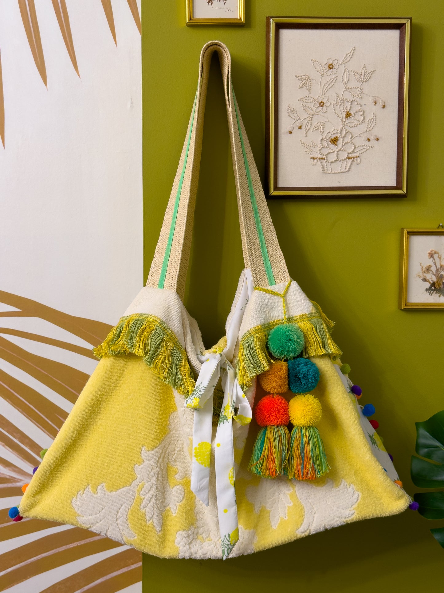 A large beach tote bag made from a vintage yellow and white towel, finished with rainbow coloured pom pom trim around the sides of the bag, the two straps are woven rattan with a lime green ribbon detail, there is a bag charm attached to one of the straps with large pom poms, one each in orange, dark teal, yellow, dark yellow and lime green with a multicolour tassel, there is a bow closure made from white with a pineapple pattern fabric that is hanging down the front of the bag.