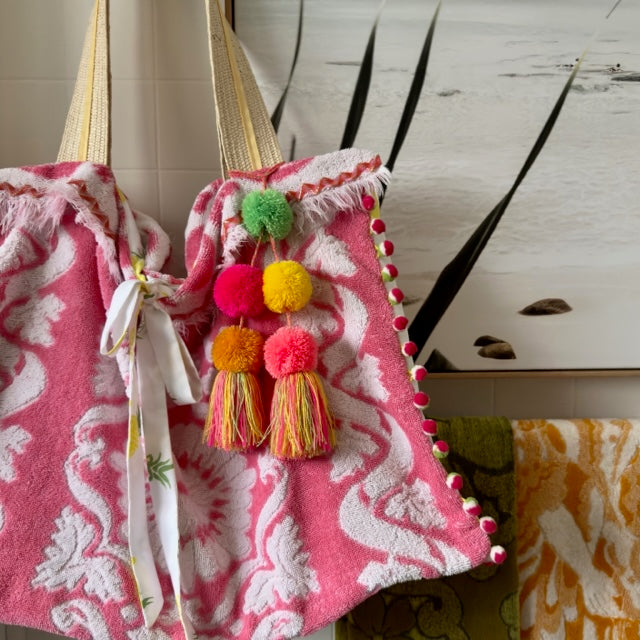 A large beach tote bag made from a vintage pink and white towel, finished with white, pink and green pom pom trim around the sides of the bag, the two straps are woven rattan with a pale yellow ribbon detail, there is a bag charm attached to one of the straps with large pom poms, one each in pink, coral, yellow, dark yellow and lime green with a multicolour tassel, there is a bow closure made from white with a pineapple pattern fabric that is hanging down the front of the bag.